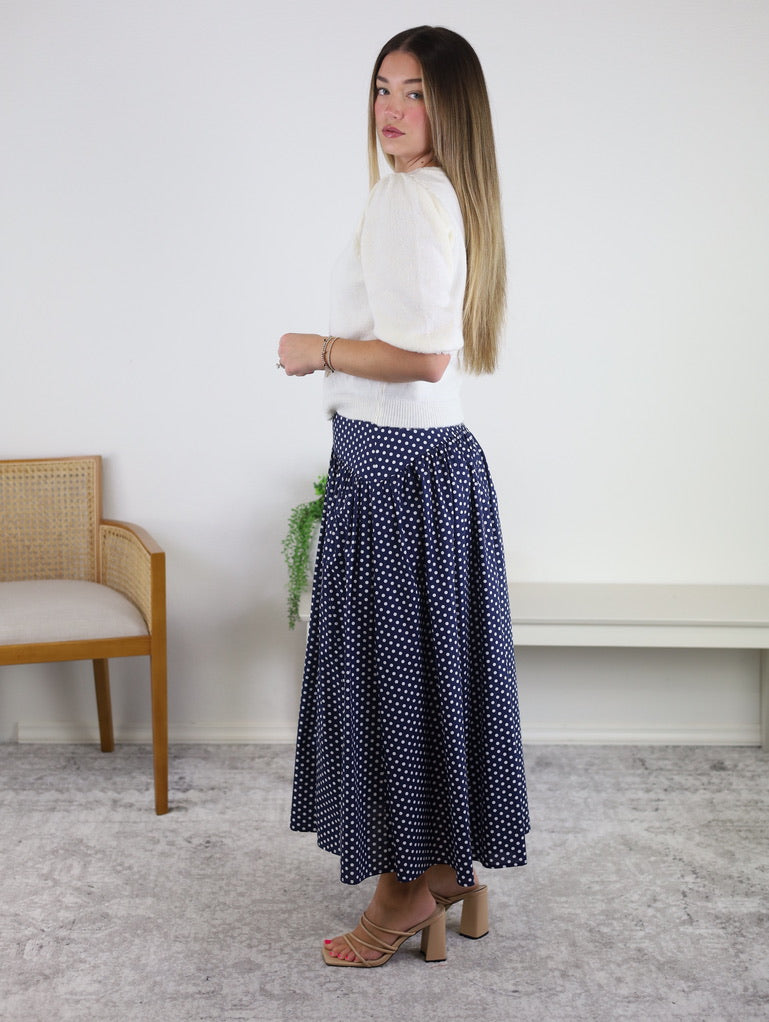 A woman stands sideways in a bright room wearing the Monica Short Sleeve Cardigan, styled with a navy blue polka dot skirt and tan heeled sandals. In the background are a wooden chair and a white bench with a plant.