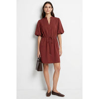 A woman wears a Naked Zebra - Puff Sleeve Cinched Waist Mini Dress: Navy / L in a rust color with a v-neckline, puff sleeves, and a drawstring waist, paired with pointed-toe flats and a small handbag.