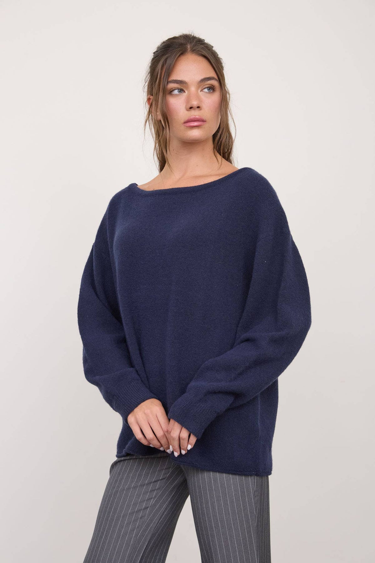 A woman wears The Workshop Studio - BOAT NECK OVERSIZED KNIT PULLOVER SWEATER in Red Wood and gray pinstriped pants, standing against a plain light background while looking slightly to the right with her hands loosely clasped.