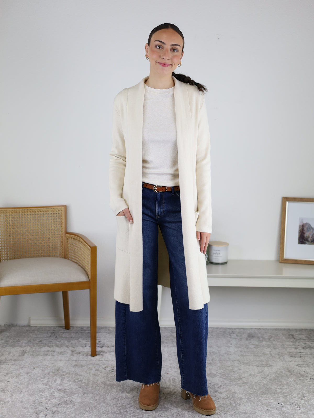 A woman stands indoors wearing the Parkland Long Cardigan in cream, styled over a light top with blue wide-leg jeans and tan shoes. She is positioned near a chair and a bench that holds a framed photo and candle. The minimalist, neutral background perfectly highlights this effortlessly chic fall look.