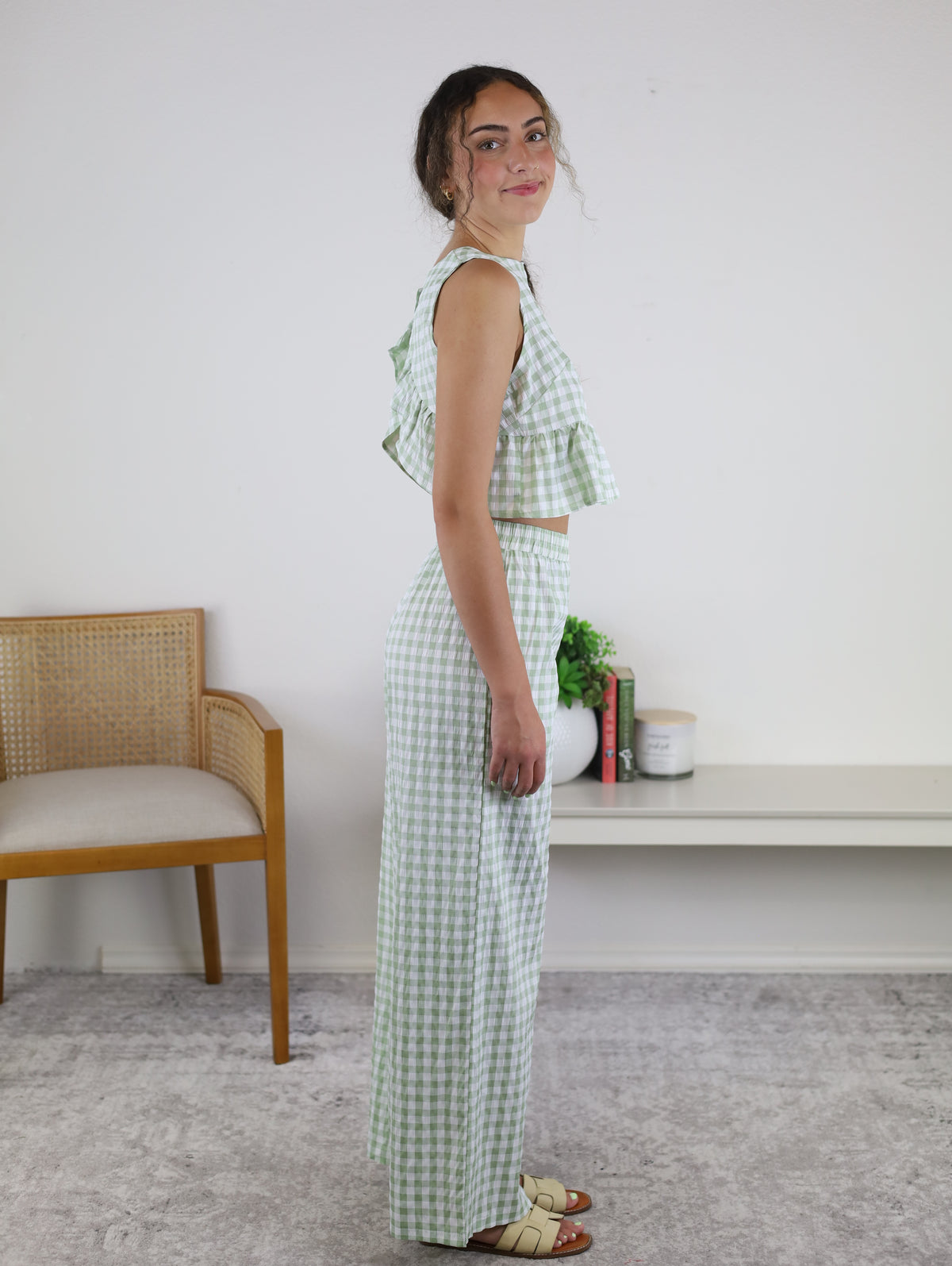 A woman stands sideways wearing the Myra Gingham Print Ruffled Set in sage, which features a sleeveless crop top and matching high-waisted pants. She is indoors, with a wooden chair, a small shelf, and a plant visible in the background.