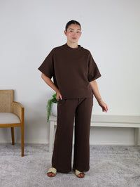 A woman stands indoors wearing the Grover Lounge Set, a loose-fitting, dark brown ensemble featuring a matching top and wide-leg pants. She pairs the set with beige sandals and is posed in a minimal room with a white wall, a bench, a plant, and a wooden chair.