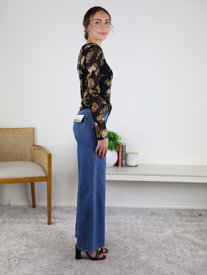 A woman is smiling and holding a phone as she stands sideways in a room with a chair, shelf, books, and a plant. She is wearing the Nightfall Rose Floral Mesh Asymmetrical Top, featuring sheer mesh sleeves, paired with blue wide-leg jeans and heeled sandals.