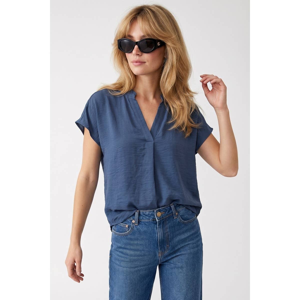A woman wearing a Naked Zebra - Drop Shoulder Regular Blouse: Chocolate / L in a blue drop shoulder woven polyester v-neck style with blue jeans.