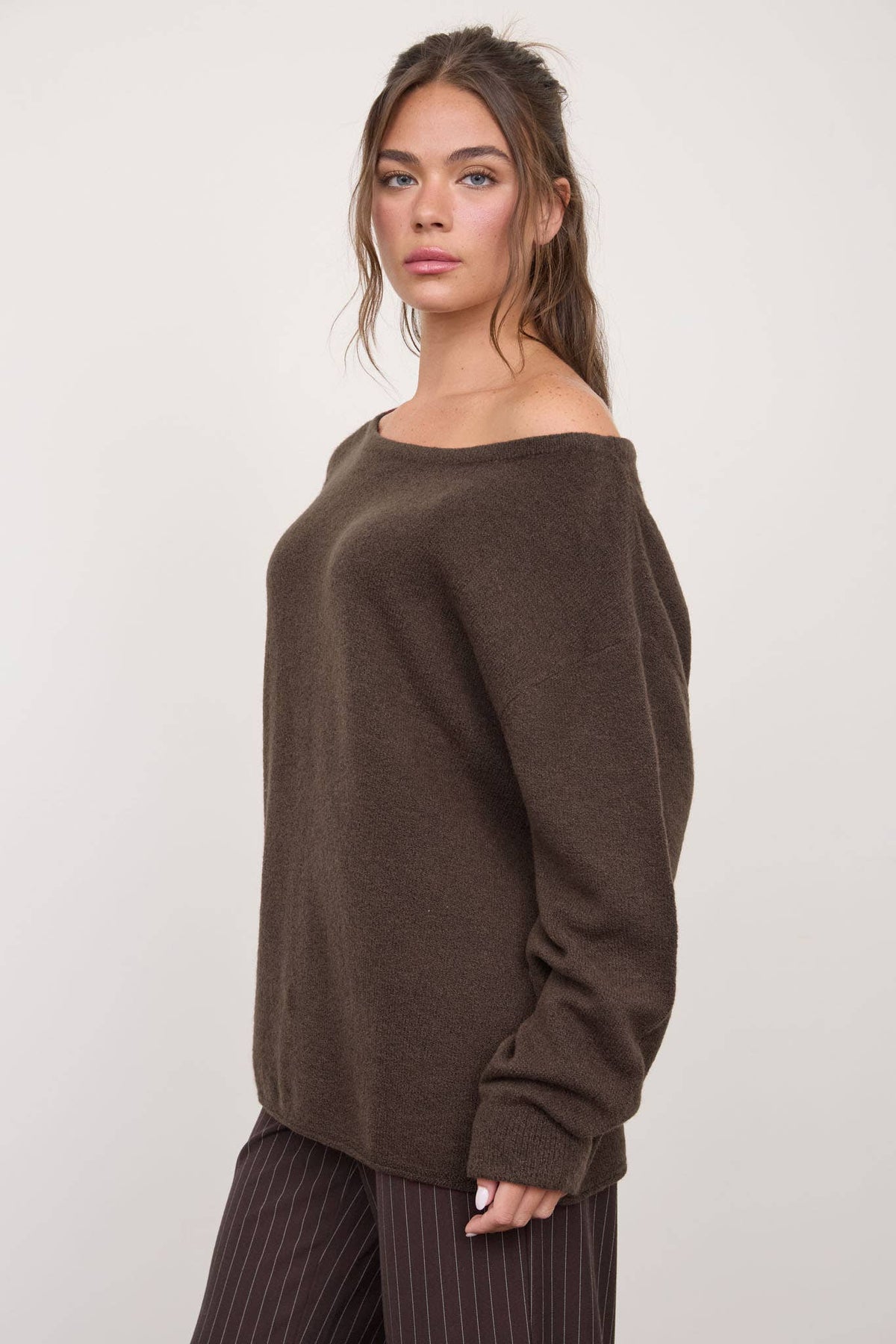 A woman with long brown hair stands against a plain light background wearing The Workshop Studio’s Boat Neck Oversized Knit Pullover Sweater in Red Wood, paired with pinstriped brown pants. The relaxed silhouette of the sweater adds a stylish touch to her outfit.