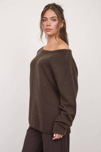A woman with long brown hair stands against a plain light background, wearing The Workshop Studio’s Boat Neck Oversized Knit Pullover Sweater in Oatmeal and striped dark pants. With its relaxed, oversized fit and boat neckline, the sweater enhances her effortless style as she looks at the camera with a neutral expression.