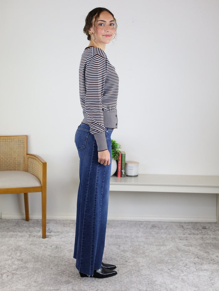 A woman stands sideways indoors, wearing the Quinn Striped Cardigan layered over a striped long-sleeve shirt, paired with wide-leg blue jeans and black shoes. In the background, there is a wooden chair, a bench with books, and a plant.