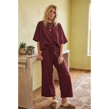 A woman stands indoors in a sunlit room with yellow-green walls and a patterned rug, wearing the Heather Brushed Stretch Boxy Top & Wide Leg Pant Set and slippers, leaning on a wooden table with potted plants.