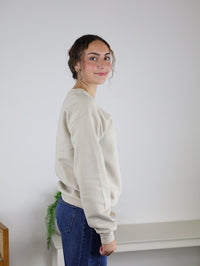 A person with curly hair stands sideways in a simple room with neutral decor and a touch of greenery, smiling slightly while wearing the Homebody Sweatshirt—a relaxed fit, light beige unisex piece crafted from a soft cotton polyester blend—paired with blue jeans.