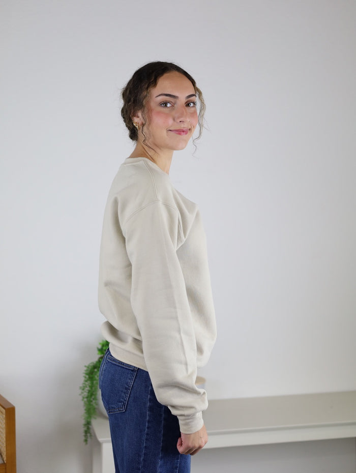 A person with curly hair stands sideways in a simple room with neutral decor and a touch of greenery, smiling slightly while wearing the Homebody Sweatshirt—a relaxed fit, light beige unisex piece crafted from a soft cotton polyester blend—paired with blue jeans.