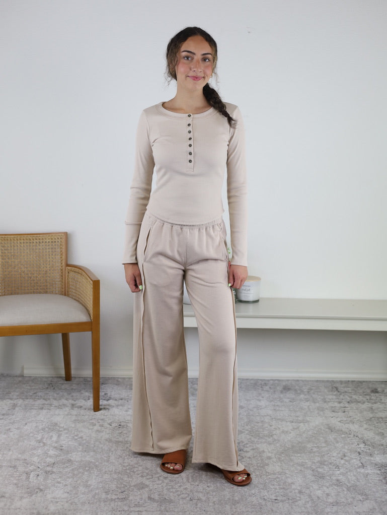 A woman stands indoors on a light carpet, smiling as she wears the Statler Lounge Set—a beige ensemble with a henley button-up top and matching wide-leg pants. She pairs the outfit with brown sandals and has her hair in a side braid. A wooden chair and white table are visible in the background.