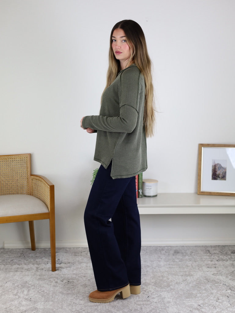 A woman with long hair stands sideways wearing the Finley Sweater, dark pants, and tan platform shoes in a minimal room with a wooden chair, bench, decor accents, and a framed photo on the wall.