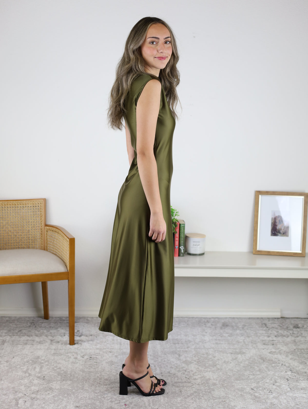 A woman in profile wears a sleeveless olive green Desi Drape Neckline Maxi Dress with a draped neckline and black heeled sandals.