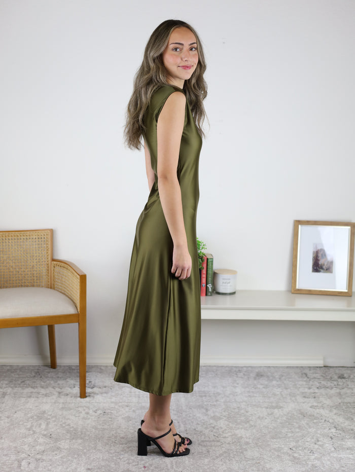 A woman in profile wears a sleeveless olive green Desi Drape Neckline Maxi Dress with a draped neckline and black heeled sandals.