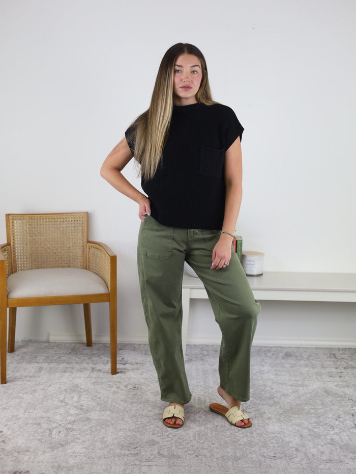 A woman with long hair wears a black short-sleeve top, the Josie Stretch Twill Utility Barrel jeans in olive green, and white sandals, standing indoors near a wooden chair and a white table against a white wall.