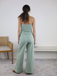 A woman with curly hair stands facing away, wearing the Kia Striped Tube Top and Pants Set in a mint stripe pattern. She is near a wooden chair and a white console table against a white wall.
