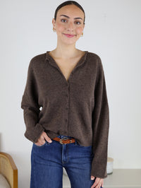 A woman with dark hair tied back wears the Beth Lightweight Cardigan in a relaxed fit, paired with blue jeans and a belt. She stands indoors, smiling with one hand in her pocket, against a plain white background.