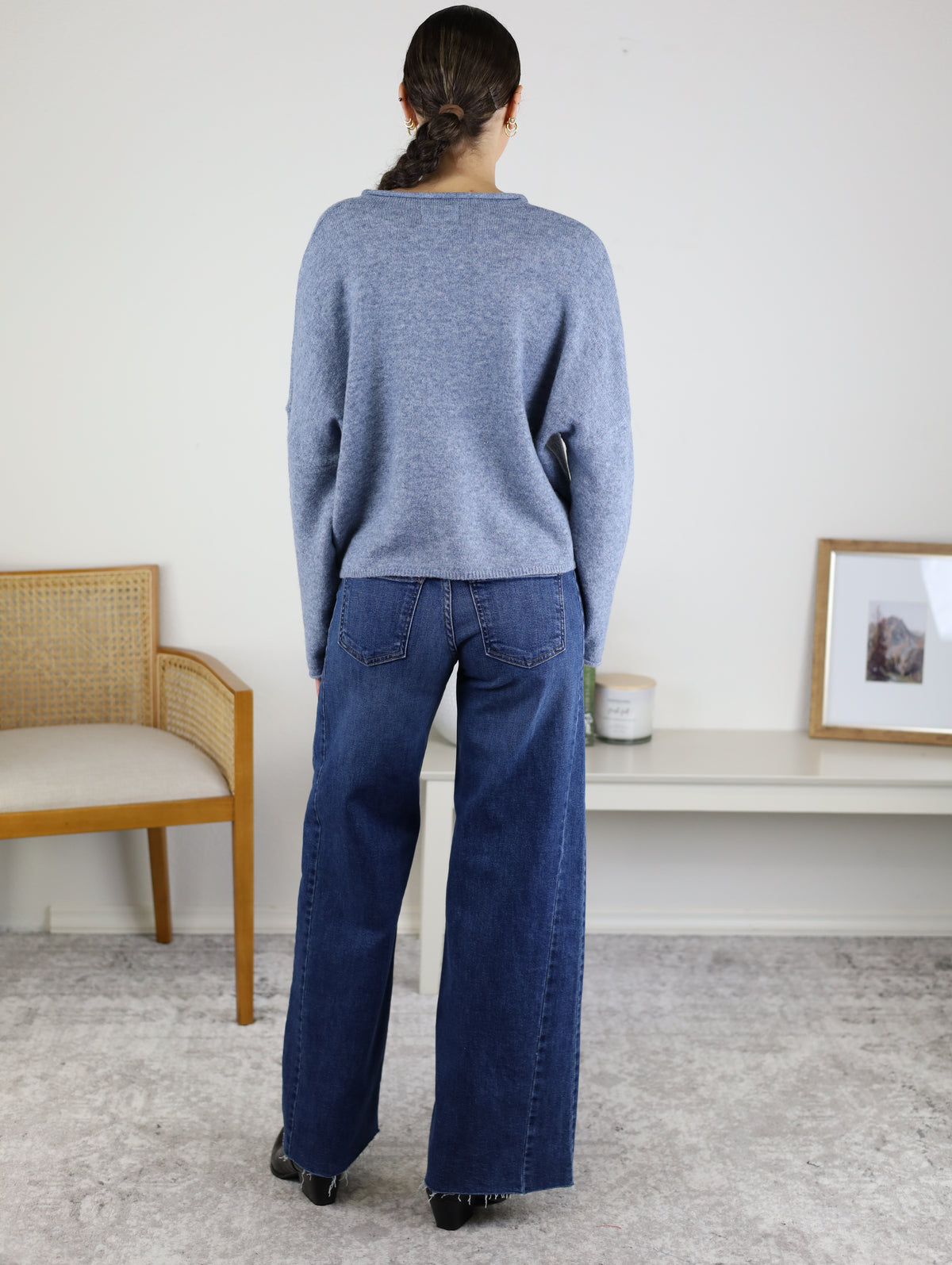 A person with dark hair in a ponytail stands facing away, wearing the Beth Lightweight Cardigan and wide-leg jeans. There is a wooden chair and a white console table with decor in a light room.