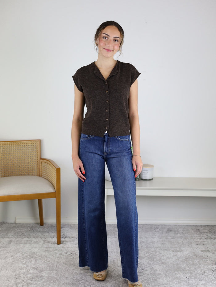 A woman stands indoors against a white wall, smiling as she wears the Camden Lightweight Cardigan layered over a short-sleeve brown button-up top and paired with blue wide-leg jeans. A chair and shelf are visible in the background.