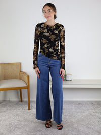 A woman stands indoors, smiling with her hands relaxed by her sides, wearing the Nightfall Rose Floral Mesh Asymmetrical Top featuring sheer mesh sleeves, paired with wide-leg blue jeans and black sandals. A chair and candles are visible in the background.