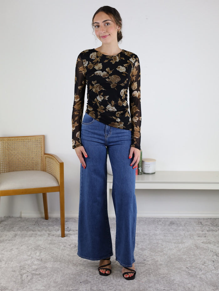A woman stands indoors, smiling with her hands relaxed by her sides, wearing the Nightfall Rose Floral Mesh Asymmetrical Top featuring sheer mesh sleeves, paired with wide-leg blue jeans and black sandals. A chair and candles are visible in the background.