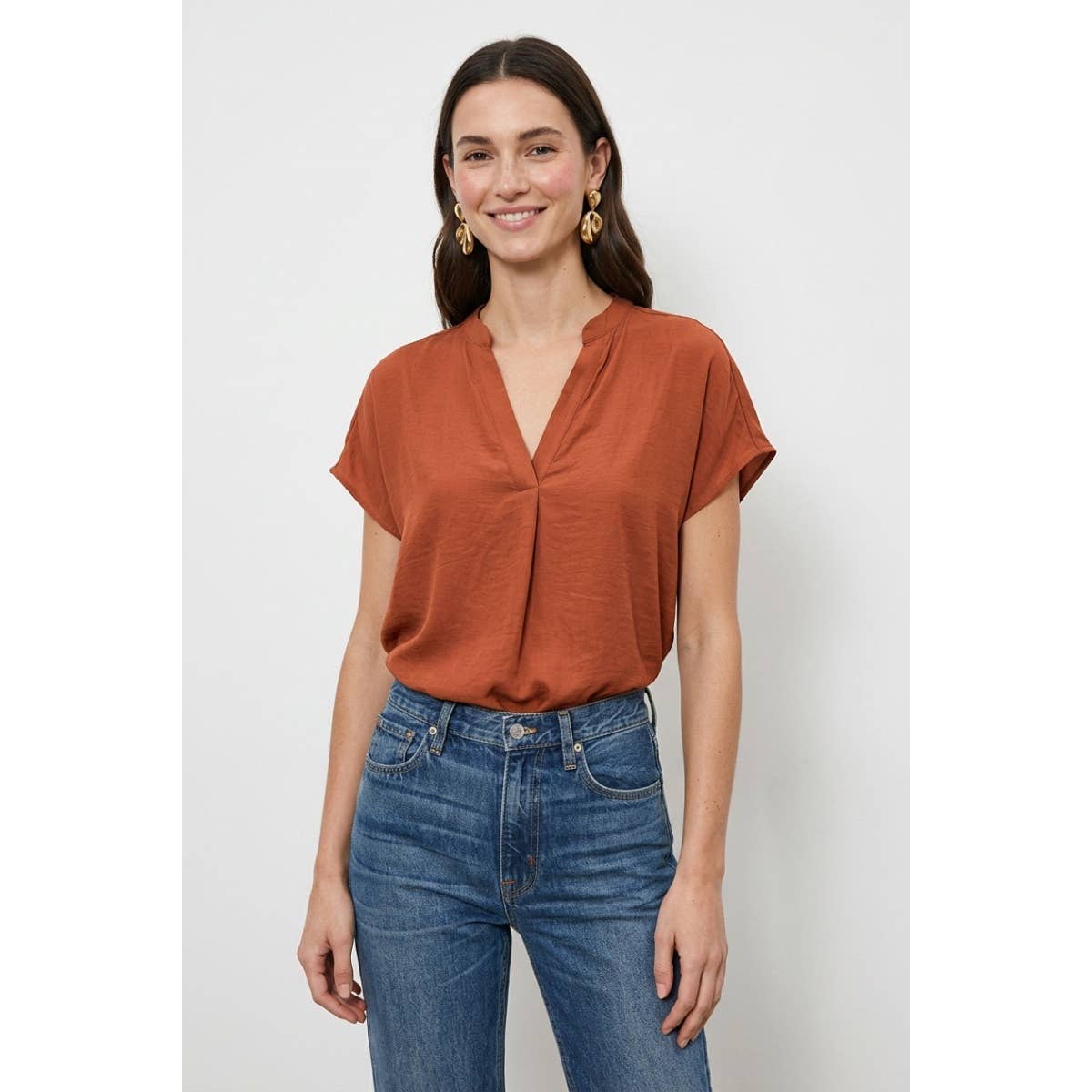 A woman wears a rust-colored Naked Zebra - Drop Shoulder Regular Blouse: Chocolate / L with a v-neckline and drop shoulders tucked into blue jeans.
