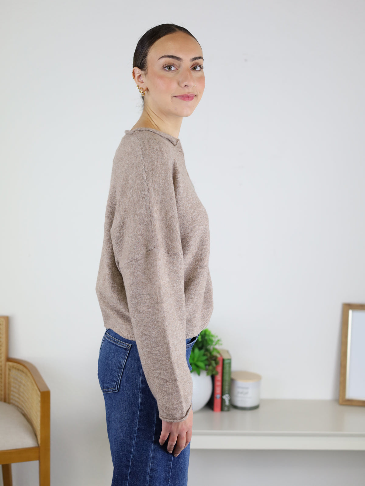 A woman with dark hair in a bun wears the Beth Lightweight Cardigan in beige and blue jeans, standing sideways in a bright room with a table, books, plant, and a framed object in the background.