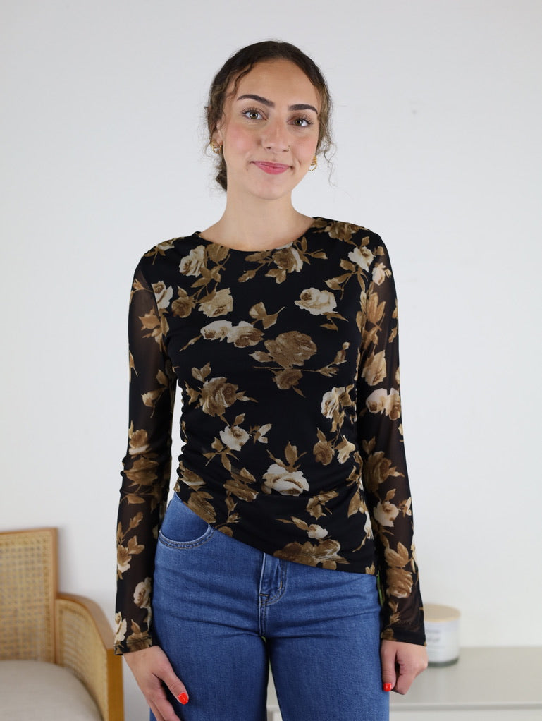 A woman wearing the Nightfall Rose Floral Mesh Asymmetrical Top with sheer mesh sleeves and blue jeans stands indoors, smiling softly at the camera.