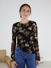 A woman wearing the Nightfall Rose Floral Mesh Asymmetrical Top with sheer mesh sleeves and blue jeans stands indoors, smiling softly at the camera.