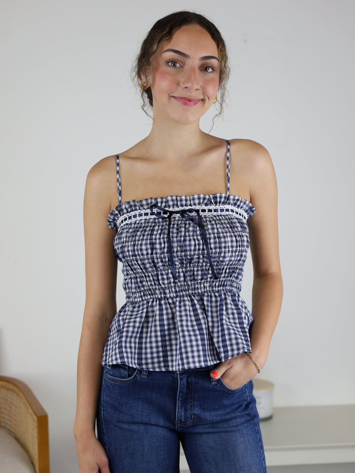 A person with wavy hair smiles while wearing the Lonnie Smocked Gingham Peplum Top—a blue-and-white checkered piece with adjustable spaghetti straps and ruffle trim—paired with blue jeans, standing indoors against a white wall and furniture background.