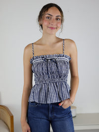 A person with wavy hair smiles while wearing the Lonnie Smocked Gingham Peplum Top—a blue-and-white checkered piece with adjustable spaghetti straps and ruffle trim—paired with blue jeans, standing indoors against a white wall and furniture background.