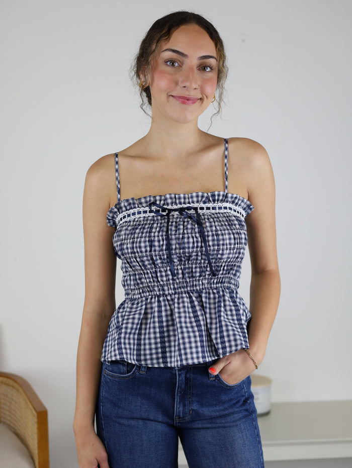 A person with wavy hair smiles while wearing the Lonnie Smocked Gingham Peplum Top—a blue-and-white checkered piece with adjustable spaghetti straps and ruffle trim—paired with blue jeans, standing indoors against a white wall and furniture background.