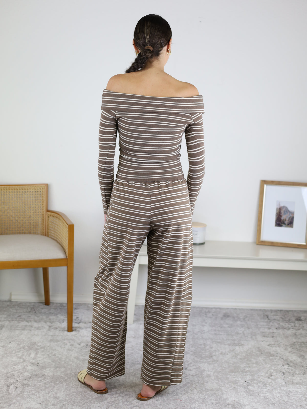 Wearing the Linn Off Shoulder Top Barrel Pant Set in brown and white stripes, a woman stands facing away in a minimal room furnished with a chair, desk, framed photo, and neutral décor—perfectly capturing an effortless loungewear look.