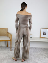 Wearing the Linn Off Shoulder Top Barrel Pant Set in brown and white stripes, a woman stands facing away in a minimal room furnished with a chair, desk, framed photo, and neutral décor—perfectly capturing an effortless loungewear look.