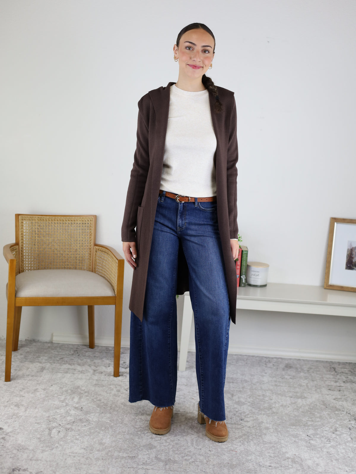 A woman stands indoors wearing the Pinch Me Long Cardigan layered over a cream sweater, paired with blue wide-leg jeans and brown shoes for an effortlessly cozy look. In the background, a wooden chair and a low table decorated with decor items add to the inviting setting.