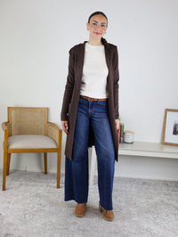 A woman stands indoors wearing the Pinch Me Long Cardigan layered over a cream sweater, paired with blue wide-leg jeans and brown shoes for an effortlessly cozy look. In the background, a wooden chair and a low table decorated with decor items add to the inviting setting.