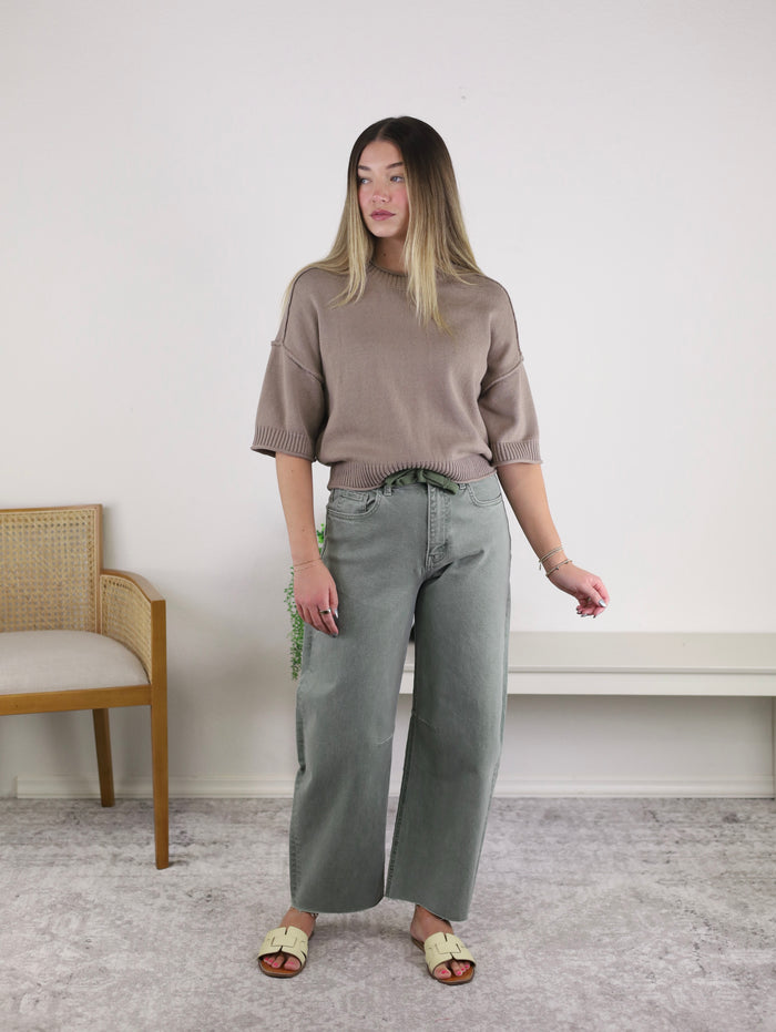A woman stands indoors wearing a taupe sweater, cream sandals, and the Willy High Rise Barrel Jean in sage green with a curved barrel leg silhouette. A wooden chair and a white bench are in the background on a light gray carpet.