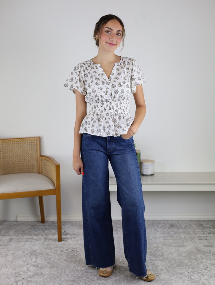Wearing the Tovar Floral Blouse, a woman stands indoors, smiling with one hand in her pocket. Crafted from 100% cotton, this short-sleeved peplum top delivers comfort and effortless style. She pairs it with wide-leg jeans while a wooden chair and shelf appear in the background.