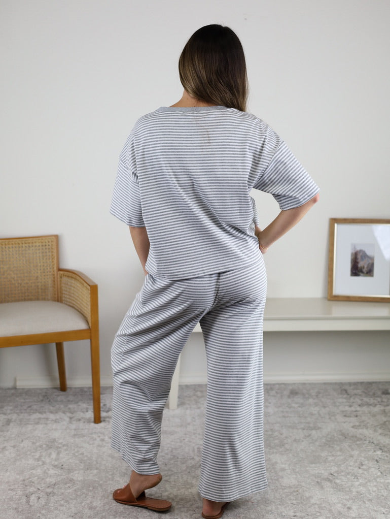 A person with long hair, wearing the Alanna Striped Knit Top and Pants Set in light gray and white, stands indoors near a chair, shelf, and framed picture on a white wall.