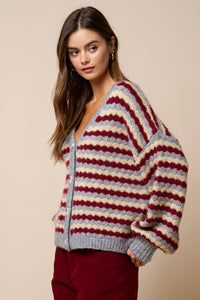 A woman stands against a beige background, looking to the side with a neutral expression while wearing the Lalavon Relaxed Stripe V-Neck Button Cardigan in grey, burgundy, and beige (size L), featuring a loose fit and long sleeves.