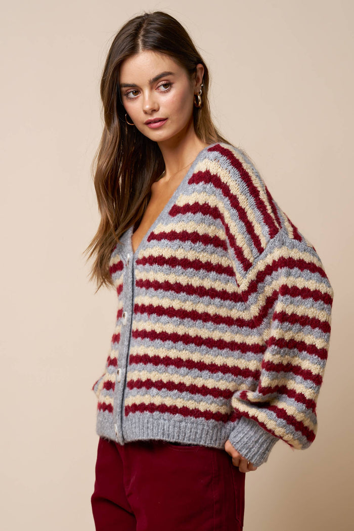 A woman stands against a beige background, looking to the side with a neutral expression while wearing the Lalavon Relaxed Stripe V-Neck Button Cardigan in grey, burgundy, and beige (size L), featuring a loose fit and long sleeves.
