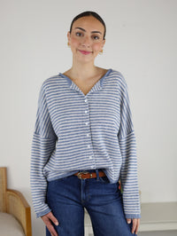 A person stands indoors, smiling, wearing the Maren Striped Cardigan—a blue and white, relaxed-fit piece—paired with classic blue jeans. The wool-blend knit of the Maren Striped Cardigan adds subtle texture to the outfit. The background is plain, with part of a chair visible on the left.