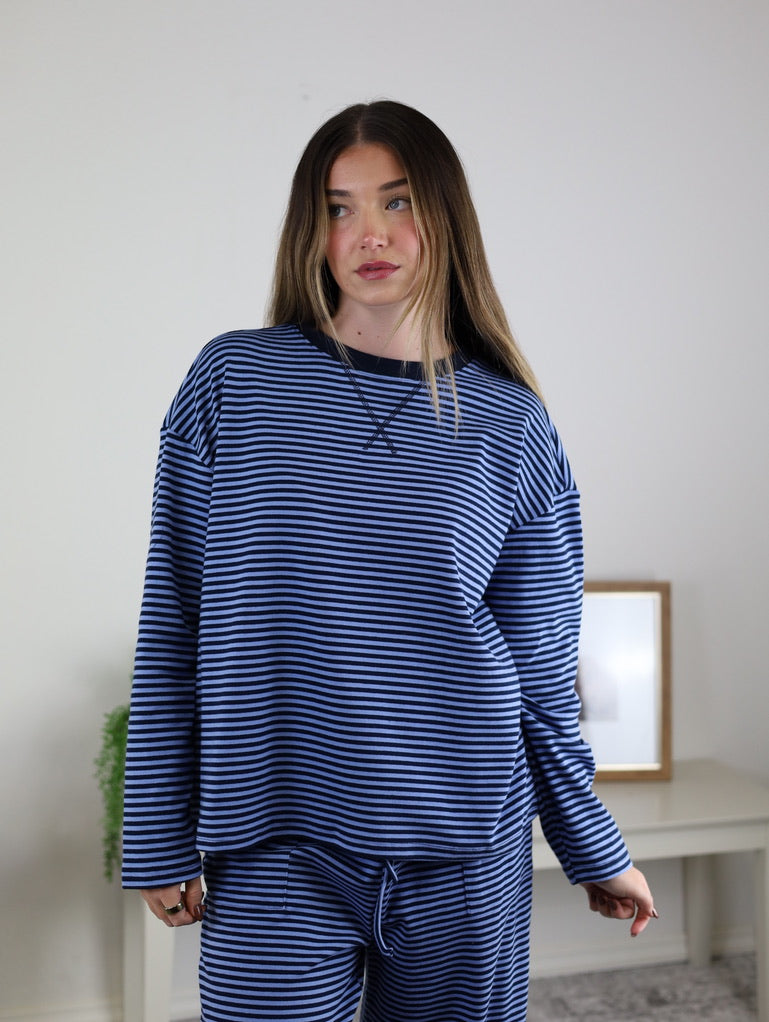 A woman wears the Misty Striped Relaxed Top & Wide Leg Pants in blue and black, standing indoors with a framed photo and green plant in the background, looking slightly to the side.
