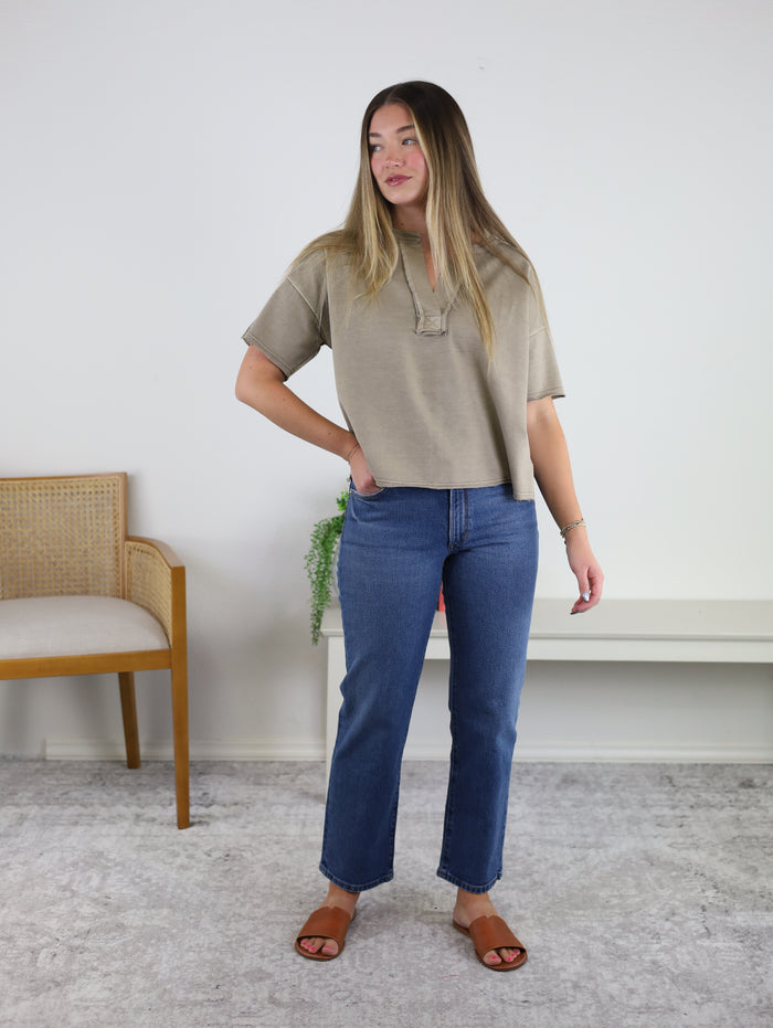A woman stands indoors wearing the Wilder Split Neck Top in taupe, featuring raw seam details, paired with blue jeans and brown sandals. She is next to a wooden chair and a bench with a potted plant, in a room with white walls and a light carpet.