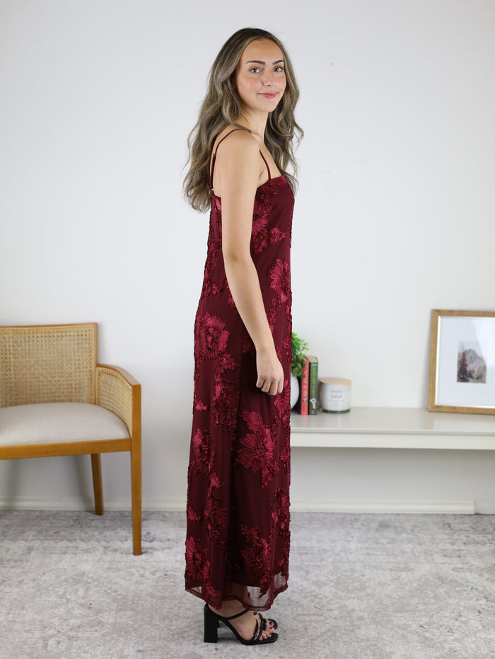 A woman wearing an Axel Applique Mesh Maxi Dress, a sleeveless, ankle-length maroon mesh gown with romantic floral applique and black heeled sandals.