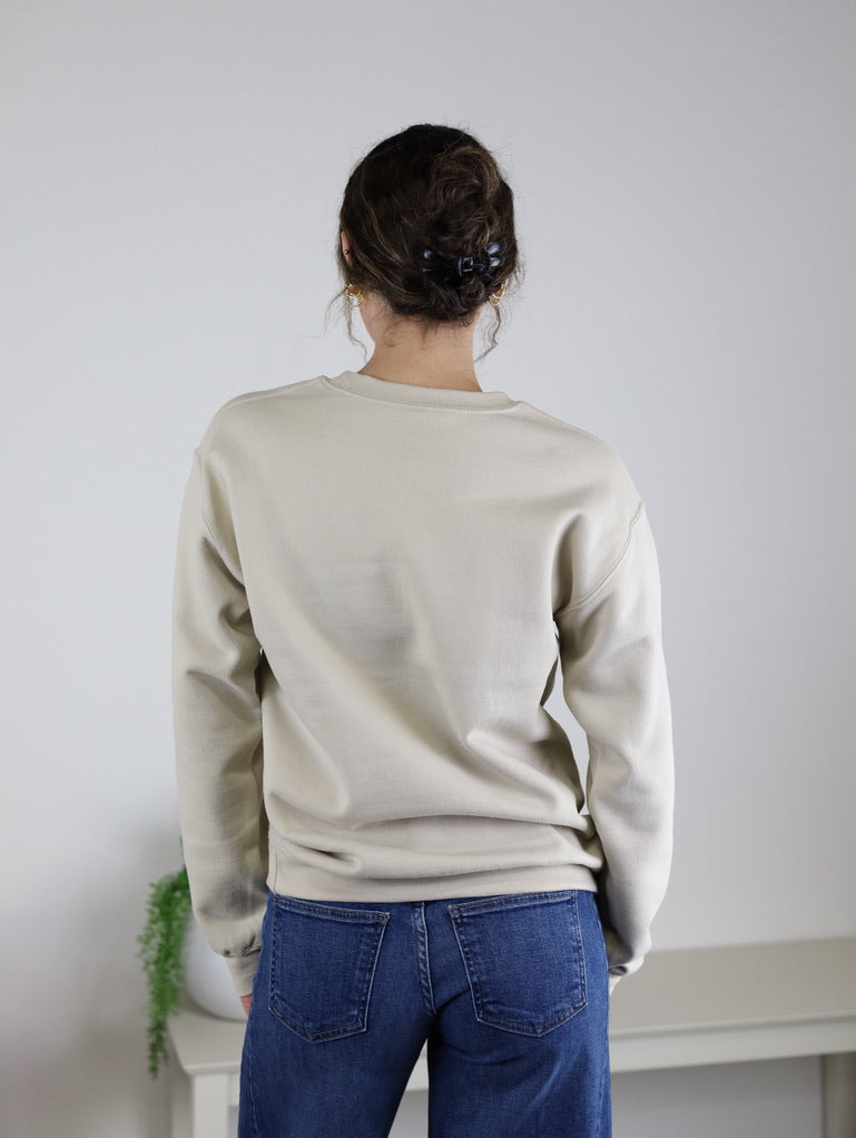 A person with curly hair in a clip is seen from behind, standing in front of a white wall and a small table with a plant, wearing the Homebody Sweatshirt—a beige unisex sweatshirt with a relaxed fit made from a comfortable cotton polyester blend—paired with blue jeans. This versatile piece is perfect for everyday style.