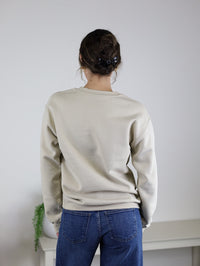 A person with curly hair in a clip is seen from behind, standing in front of a white wall and a small table with a plant, wearing the Homebody Sweatshirt—a beige unisex sweatshirt with a relaxed fit made from a comfortable cotton polyester blend—paired with blue jeans. This versatile piece is perfect for everyday style.
