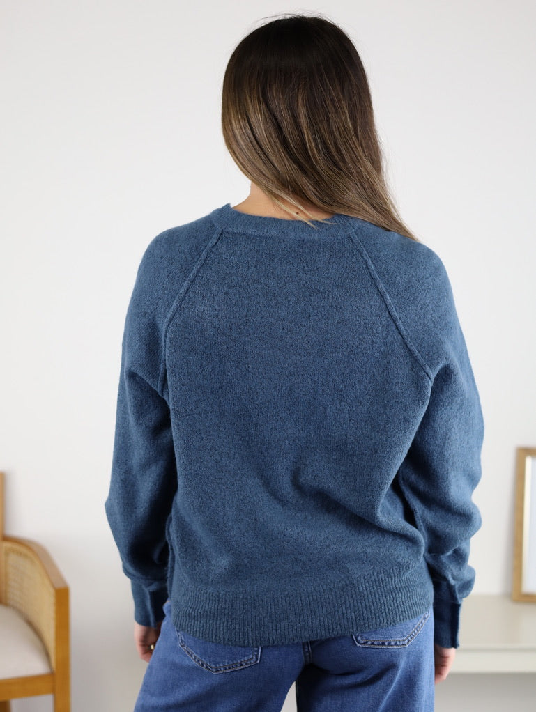 A woman with long brown hair wears the Theo Sweater and blue jeans, standing with her back to the camera in a minimal indoor setting.