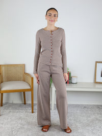 A woman stands indoors wearing the Melissa Lounge Stripe Button Down Set, which features a taupe, striped long-sleeve button down top paired with matching wide leg pants. She is smiling, with a chair and a bench adorned with decor items in the background.