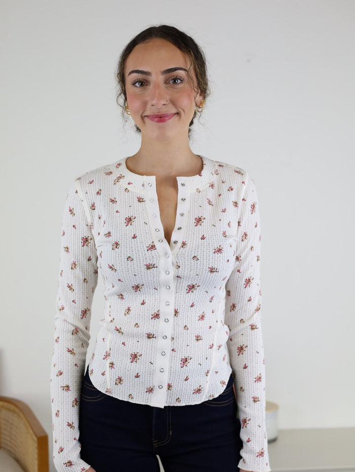 A woman with curly hair, wearing the Izzy Floral Waffle Knit Top—a white, long-sleeved floral waffle knit—and dark jeans, stands and smiles in front of a plain white wall. A small part of a chair and table are visible in the background.